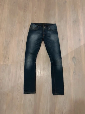Nudie jeans - Säljer ett par mörkblå jeans med snygga tvättade detaljer. Klassisk femficksmodell med kontrastsömmar och smal passform hela vägen ner. Jeansen har en enkel look som passar till det mesta och är tillverkade i stretchig denim för extra komfort.