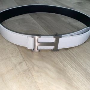 Vitt bälte med H-spänne - Snyggt vitt bälte med ikoniskt H-format metallspänne i silver. Bältet har en stilren och modern look, perfekt för att lyfta vilken outfit som helst. Tillverkat i slätt skinn med svart insida för en exklusiv känsla.