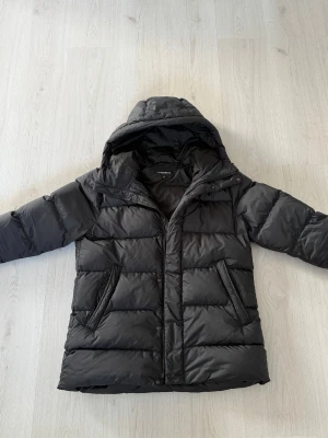 Svart dunjacka från J.Lindeberg - Snygg svart dunjacka från J.Lindeberg med huva och dragkedja framtill. Jackan har två snedställda fickor med dragkedja och diskret logga på ärmen. Perfekt för kalla vinterdagar och riktigt skön att bära. Storlek M men passar nog L med. Går för  runt 3800 som ny 