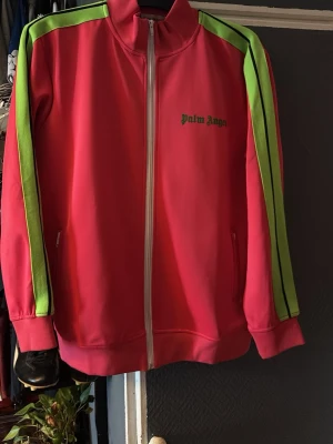 Palm Angels rosa track jacket - Säljer en snygg rosa track jacket från Palm Angels med limegröna ränder längs ärmarna och dragkedja framtill. Jackan har hög krage, två fickor och logga på bröstet. Perfekt för dig som gillar streetwear och vill sticka ut. Priset kan sönkas vid snabb affär 