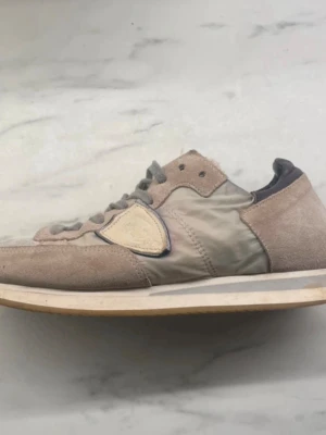 Beige sneakers från Philippe Model - Snygga beige sneakers från Philippe Model med gråa skosnören och ikonisk sköldlogga på sidan. Ovandelen är i mocka och textil, med detaljer i vitt och grått. Klassisk låg modell med vit sula och retrovibbar.