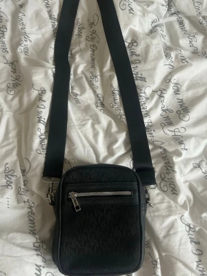 Svart axelväska från MCM - Snygg svart axelväska från MCM med justerbar bred axelrem och silverfärgade detaljer. Väskan har ett mönster med MCM-loggor över hela ytan och en praktisk ytterficka med dragkedja framtill. Perfekt för att bära det viktigaste med stil. Väskan är aldrig använd bara legat i garderoben.