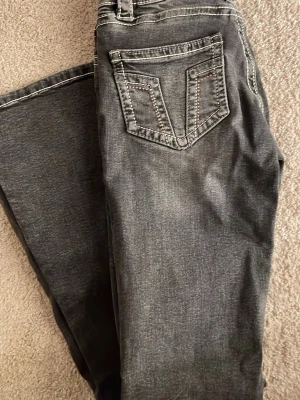 Grå jeans med kontrastsömmar - Snygga grå jeans med tydliga kontrastsömmar och en unik bakficka med V-formad detalj. Jeansen har klassisk femficksdesign och raka ben. Materialet ser ut att vara bomullsdenim med lite stretch för extra komfort.