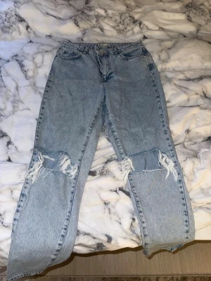 Ljusblå ripped mom jeans - Säljer ett par ljusblå mom jeans med stora slitningar över knäna. Jeansen har hög midja, raka ben och klassisk femficksmodell. Perfekta för en avslappnad och trendig look. Finns en liten fläck på dem. 