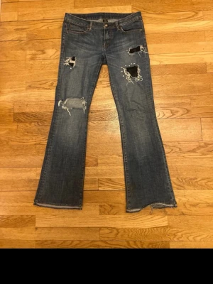 Blå slitna bootcut jeans med nitar och spets - Säljer ett par blå bootcut jeans med slitningar och hål på benen. Jeansen har coola nitar runt ett av hålen och råa kanter vid bensluten. Klassisk femficksmodell i denim med dragkedja och knapp. Perfekta för en edgy och avslappnad stil.