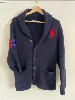 Mörkblå stickad kofta Polo Ralph Lauren - Snygg mörkblå stickad kofta från Polo Ralph Lauren med stora röda loggan på bröstet och blå ränder samt röd siffra 3 på ärmen. Koftan har knappar framtill samt 2 fickor. 