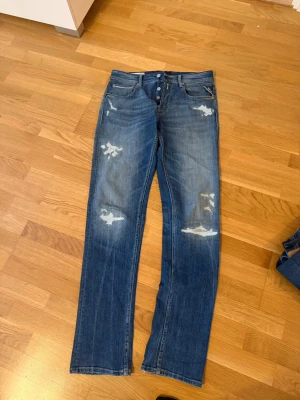 Blå straight fit jeans med slitningar - Säljer ett par blå jeans från H&M med straight fit och coola slitningar på benen. Jeansen har klassisk femficksmodell, knäppning med knappar och en avslappnad vibe. Perfekta för dig som gillar en trendig och avslappnad stil.
