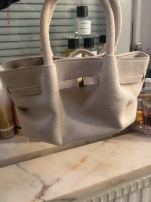 Beige mocka handväska med gulddetalj - Stilren beige handväska i mjuk mocka med två rejäla handtag och en snygg guldfärgad detalj framtill. Väskan har en klassisk, rymlig form och passar perfekt för dig som gillar minimalistisk design och neutrala färger.