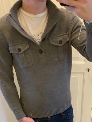 Grå stickad tröja med fickor - Snygg grå stickad tröja med två bröstfickor med lock och knappar. Tröjan har en bred krage med knäppning framtill och ribbade muddar vid ärmslut och nederkant. Perfekt för lager-på-lager och chill stil. Från s.oliver