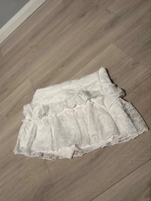 Vit spetskjol med volang och dragkedja - Söt vit kortkjol i spets med volangdetaljer och dragkedja i sidan. Kjolen har ett tunt innerfoder och ett romantiskt uttryck med sina spetslager. Perfekt för dig som gillar en feminin och trendig stil.