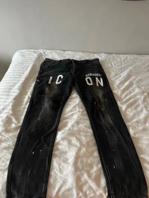 Svarta Dsquared2 ICON jeans - Säljer ett par svarta jeans från Dsquared2 med stor vit 'ICON'-text framtill. Jeansen har smal passform, klassisk femficksdesign och coola slitningar samt färgstänk för en edgy look. Perfekta för dig som vill sticka ut med streetwear-vibe.