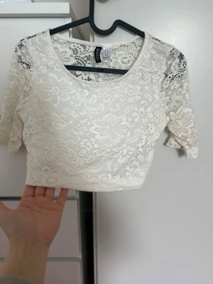 Vit spets croppad topp  - Superfin vit croppad topp från H&M i spets med blommigt mönster. Toppen har rund halsringning och korta ärmar. Perfekt att styla med höga jeans eller kjol för en trendig look.