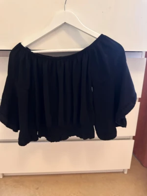 Svart offshoulder croppad topp - Trendig svart offshoulder topp med croppad passform och vida ärmar. Toppen har resår vid midjan och ärmarna för en snygg rynkad effekt. Perfekt för dig som vill ha en luftig och stilren look.