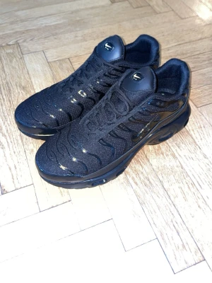 Nike TN’s  helsvarta sneakers - Nike Air Max Plus sneakers i helsvart med mesh- och syntetmaterial. Skorna har den ikoniska vågdesignen, synlig Air Max-dämpning i sulan och TN-logga på plösen. Snygga detaljer i svart lack och klassisk Nike Swoosh på sidan. Perfekta för dig som gillar streetwear.