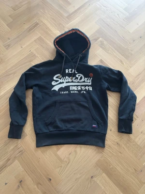 Svart/Blå Superdry hoodie med tryck - Svart/Blå hoodie från Superdry med stort vitt tryck och japanska tecken på bröstet. Klassisk känguruficka framtill, dragsko i huvan och mjukt fleecefoder. Detaljer i blått och orange på insidan av huvan. Perfekt för dig som gillar streetwear och snygga loggor.
