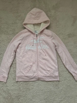 Ljusrosa hoodie från Hollister - Mysig ljusrosa hoodie från Hollister med broderad logga på bröstet. Tröjan har huva med snörning, dragkedja framtill och två fickor. Insidan är fodrad med fluffigt, vitt teddyfoder som gör den extra varm och skön.