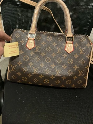 Louis Vuitton Monogram handväska - Snygg handväska från Louis Vuitton i klassiskt brunt monogrammönster med beige detaljer och röda sömmar. Väskan har guldfärgade metalldetaljer, dragkedja och två handtag. Ikonisk design som verkligen sticker ut.
