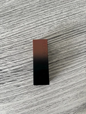 Huda Beauty Board Meeting läppstift - Snyggt matt läppstift från Huda Beauty i nyansen Board Meeting. Hylsan är fyrkantig och har en svart till brun ombre-design i plast. Perfekt för dig som gillar trendiga, neutrala toner och vill ha ett stilrent läppstift i sminkväskan.