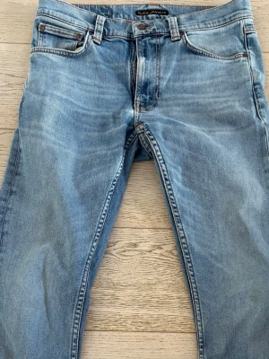 Ljusblå jeans från Nudie Jeans - Säljer ett par ljusblå jeans från Nudie Jeans med klassisk femficksmodell och raka ben. Jeansen har snygga slitningar och kontrastsömmar. Perfekta för dig som gillar en avslappnad och tidlös stil.
