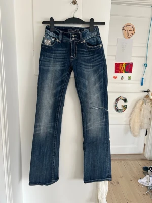 Mörkblå bootcut jeans Miss Me - Säljer ett par mörkblå bootcut jeans från Miss Me med slitningar och snygga paljettdetaljer på fickorna. Jeansen har låg midja, kontrastsömmar och en liten reva på ena benet. Jeansen är köpa på second hand, står storlek 29 inne i jeansen i US storlek tror jag, vilket jag skulle säga passar typ strlk 25,26, i vanlig storlek! Säljer då de ej kommer till användning samt är lite för små. Jeansen har några slitningar som är lagade med lappar, samt lite lösa knappar och en silverdetalj som ramlat lite