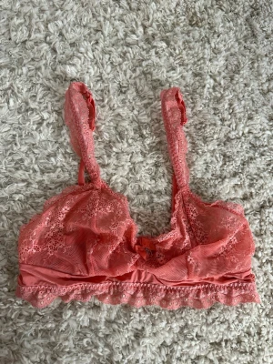 Rosa spets-bh med rosett och charm strl 38 - Söt rosa bh i spets med volangkant och dekorativ rosett framtill. Bh:n har justerbara axelband och en liten metallcharm med bokstaven 'M' vid rosetten. Materialet är genomskinlig spets och mjuk mesh för en romantisk look.