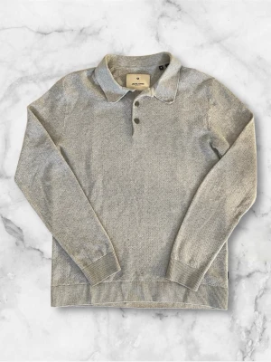 Grå stickad pikétröja från Jack and Jones - Snygg grå stickad pikétröja från Jack and Jones  med klassisk krage och tre knappar framtill. Tröjan har långa ärmar, ribbade muddar och nederkant. Perfekt för en stilren och avslappnad look.