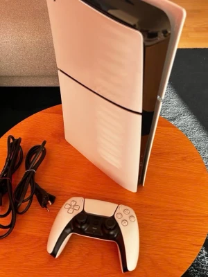 Sony PlayStation 5 med handkontroll - Säljer en PlayStation 5 från Sony i vit och svart design. Konsolen har en futuristisk, stilren form och medföljer gör en matchande DualSense-handkontroll samt strömkabel och USB-kabel. Perfekt för gaming på nästa nivå med kraftfull prestanda och modern look.