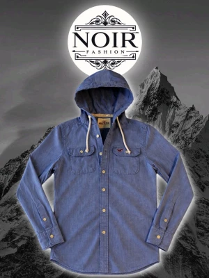 Blå huvskjorta från Hollister - Blå skjorta från Hollister med huva och vita snören. Skjortan har två bröstfickor med knapp, vita knappar framtill och på ärmsluten samt en liten broderad logga på bröstet. Perfekt för en avslappnad och cool stil.