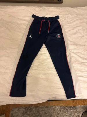 PSG x Jordan blåa mjukisbyxor - Mörkblå mjukisbyxor från Jordan i samarbete med PSG. Byxorna har vita sidopaneler, röda detaljer och dragsko i midjan. PSG-logga och Jumpman-logga på framsidan. Tillverkade i ett mjukt och sportigt material, perfekta för chill eller träning.