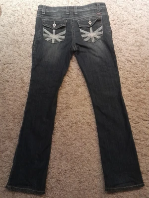 Mörkblå jeans med Union Jack-detalj - Mörkblåa Makers Of True Originals bootcut jeans för kvinnor. Ben innerlängd cirka 82cm, midja cirka 43cm halv mått (runt hela midjan är cirka 86cm)