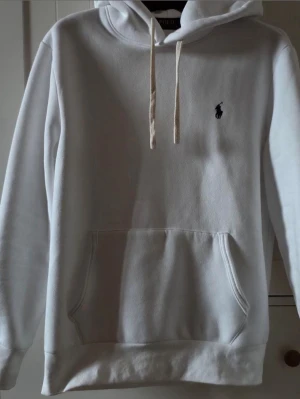 Vit hoodie från Polo Ralph Lauren - Snygg vit hoodie från Polo Ralph Lauren med klassisk huva och dragsnören. Har en stor magficka och den ikoniska broderade loggan i svart på bröstet. Tillverkad i mjukt bomullsmaterial som är skönt att ha på sig. Perfekt för en avslappnad och stilren look.