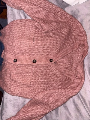 Rosa stickad kofta med knappar - Mysig rosa stickad kofta med stora bruna knappar framtill och två rejäla fickor. Koftan har en avslappnad passform, ribbade muddar och är perfekt för lager på lager. Passar dig som gillar en chill och trendig stil.