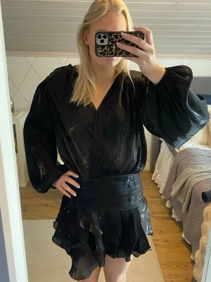 Svart omlottklänning med volang - Kort svart omlottklänning med volangdetaljer och långa puffärmar. Klänningen har en markerad midja och ett lätt glansigt tyg som ger en elegant känsla. Perfekt för dig som gillar en trendig och chic look.