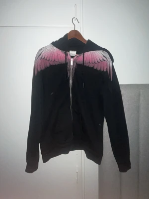 Äkta marcelo burlon kofta  - Säljer en svart hoodie från Marcelo Burlon med dragkedja och huva. Hoodien har ett fett tryck av rosa och vita vingar över axlarna och bröstet. Perfekt för dig som vill sticka ut med en unik och snygg design. Materialet är mjukt och skönt bomullstyg.