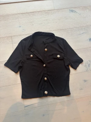 Svart kortärmad skjorta med knappar - Stilren svart skjorta med korta ärmar och dekorativa guldknappar framtill. Skjortan har två fejkade bröstfickor med knappar och en lätt ribbad struktur. Perfekt för dig som gillar en trendig och clean look.