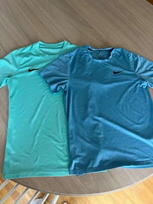 Två sporttröjor från Nike blå/turkos - Säljer två snygga sporttröjor från Nike i blått och turkost. Båda har rund hals, korta ärmar och den klassiska Nike-loggan på bröstet. Materialet är lätt och andas, perfekt för träning eller löpning. Säljer både två tillsammans. Passar M/L