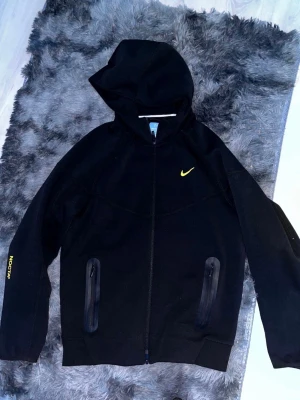 Svart Nike NOCTA hoodie med dragkedja - Snygg svart hoodie från Nike NOCTA-kollektionen med gul logga på bröstet och NOCTA-tryck på ärmen. Hoodien har dragkedja framtill, två fickor med dragkedja och en stor huva. Perfekt för en streetwear-look och riktigt bekväm passform.