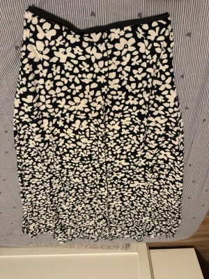 Svartvit blommig långkjol - Snygg svart långkjol med vitt blommönster och elastisk midja. Kjolen har en rak passform och är tillverkad i ett mjukt, stretchigt material som känns bekvämt mot huden. Perfekt för dig som gillar en stilren men ändå lekfull look.