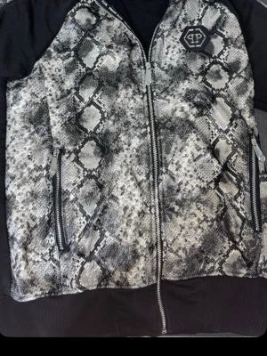 Philipp Plein hoodie med ormmönster - Snygg hoodie från Philipp Plein med ormmönstrad framsida i grått och svart, svarta ärmar och rygg med stor panda-tryck och text. Dragkedja framtill och två fickor med dragkedja. Märkeslogga på bröstet och detaljerade broderier.