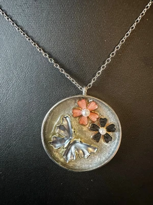 Silverfärgat halsband med fjäril och blommor - Unikt halsband med rund silverfärgad berlock som har en metallisk fjäril och två blommor, en rosa och en svart, båda med pärldetaljer i mitten. Kedjan är tunn och silverfärgad. Perfekt för dig som gillar naturinspirerade smycken och vill sticka ut.
