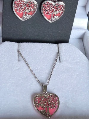 Halsband & örhängen hjärta träd motiv - Set med halsband och örhängen i form av hjärtan med trädmotiv. Smyckena är i silverfärgad metall, kedja & krokar i rostfritt stål, med rosa bakgrund och har ett unikt mönster. De lyser i mörkret med lila sken, vilket ger en cool effekt. Perfekt för dig som gillar detaljerade och personliga accessoarer. Effekten aktiveras av vanligt dagsljus, så bär det som ett vanligt smycke så sköter sig resten själv! Tål vatten, lång livslängd & skenet varar i flera timmar. Handgjort.