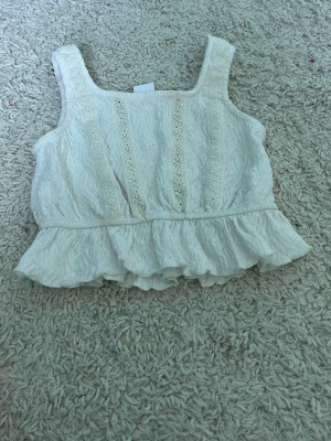 Vit volang topp med spetsdetaljer - Söt vit topp med breda axelband, volangkant nedtill och dekorativa spetsdetaljer framtill. Toppen har en lätt skrynklig struktur och är perfekt för varma dagar. Passar dig som gillar romantisk och bohemisk stil.