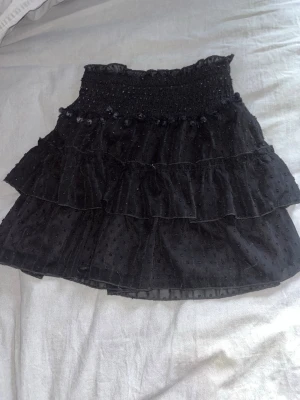 Svart volangkjol från SHEIN - Supersöt svart kortkjol från SHEIN med flera lager volanger och smock i midjan. Kjolen har diskreta prickar i tyget och små bollar längst.