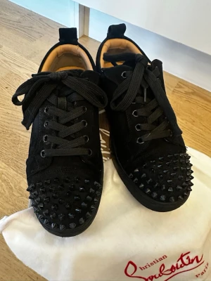 Svarta spikes-sneakers Louboutin - Svarta sneakers från Christian Louboutin med mattsvarta nitar över tån och svart mocka. Klassisk låg modell med svarta skosnören och detaljer i läder. Insidan är beige och sulan är svart. Grymt statement-plagg för dig som gillar edgy stil.