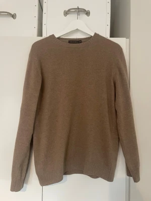 Beige stickad tröja från Oscar Jacobsson - Klassisk beige stickad tröja från Oscar Jacobsson med rund halsringning och ribbade muddar vid ärmslut och nederkant. Tröjan har en enkel och stilren design, perfekt för lager på lager och passar till det mesta. Materialet ser ut att vara mjukt och bekvämt.                                                     Kontakta gärna vid fler frågor eller bilder!