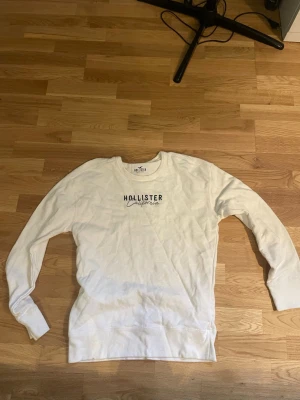 Vit sweatshirt från Hollister - Vit sweatshirt från Hollister med rund halsringning och tryckt logga på bröstet. Tröjan har långa ärmar och en klassisk passform som passar till det mesta. Perfekt för dig som gillar enkel och stilren design.