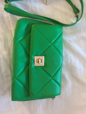 Grön quiltad handväska med axelrem - Snygg grön handväska i quiltad design med justerbar axelrem och guldfärgat vridlås. Väskan har en rektangulär form och är tillverkad i ett mjukt, läderliknande material. Perfekt för dig som vill sticka ut med en färgglad accessoar.