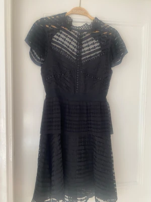 Svart spetsklänning med kort ärm - Snygg svart klänning med spetsdetaljer och korta ärmar. Klänningen har v-ringad illusion upptill, markerad midja och volangliknande kjol. Genombrutna partier och dekorativa knappar framtill ger en elegant look. Perfekt för dig som gillar detaljerad design.