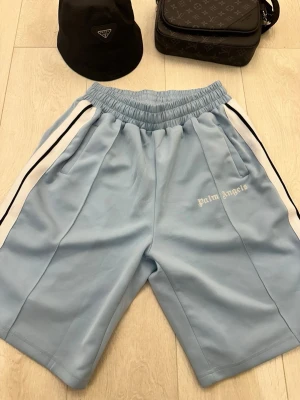 Palm Angels ljusblå track shorts - Palm Angels ljusblå track shorts med vita och svarta ränder längs sidorna. Shortsen har elastisk midja, två sidofickor och Palm Angels-logga broderad i vitt på vänster ben. Tillverkade i ett mjukt, sportigt material som ger en avslappnad vibe.