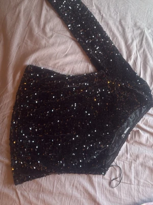 Svart paljettopp oneshoulder NA-KD - Säljer en svart oneshoulder topp från veramoda (pices)täckt med glittriga paljetter. Toppen har lång ärm på ena sidan och är axelbandslös på andra, vilket ger en unik look. Perfekt för dig som vill sticka ut och glänsa lite extra.